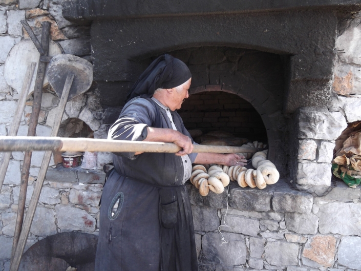 Woman in traditional coat baking bread in outside communal oven.